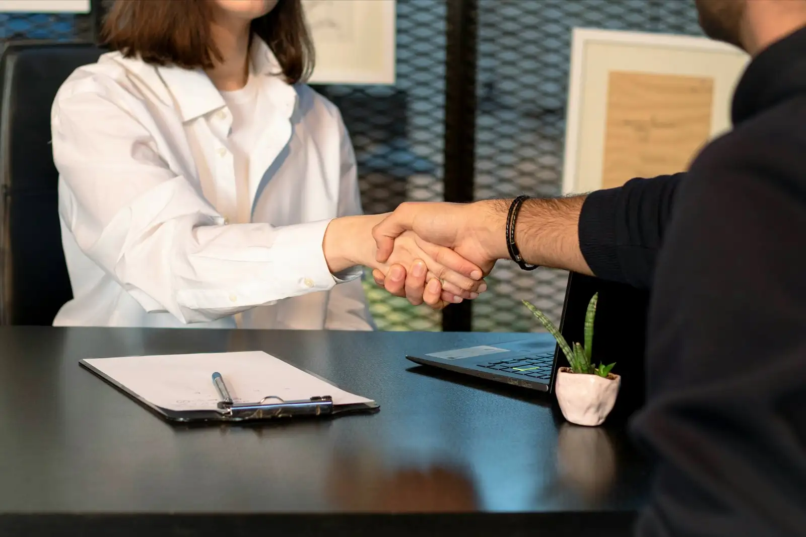 A handshake across a desk over a signed application, with the plans and laptop still open
