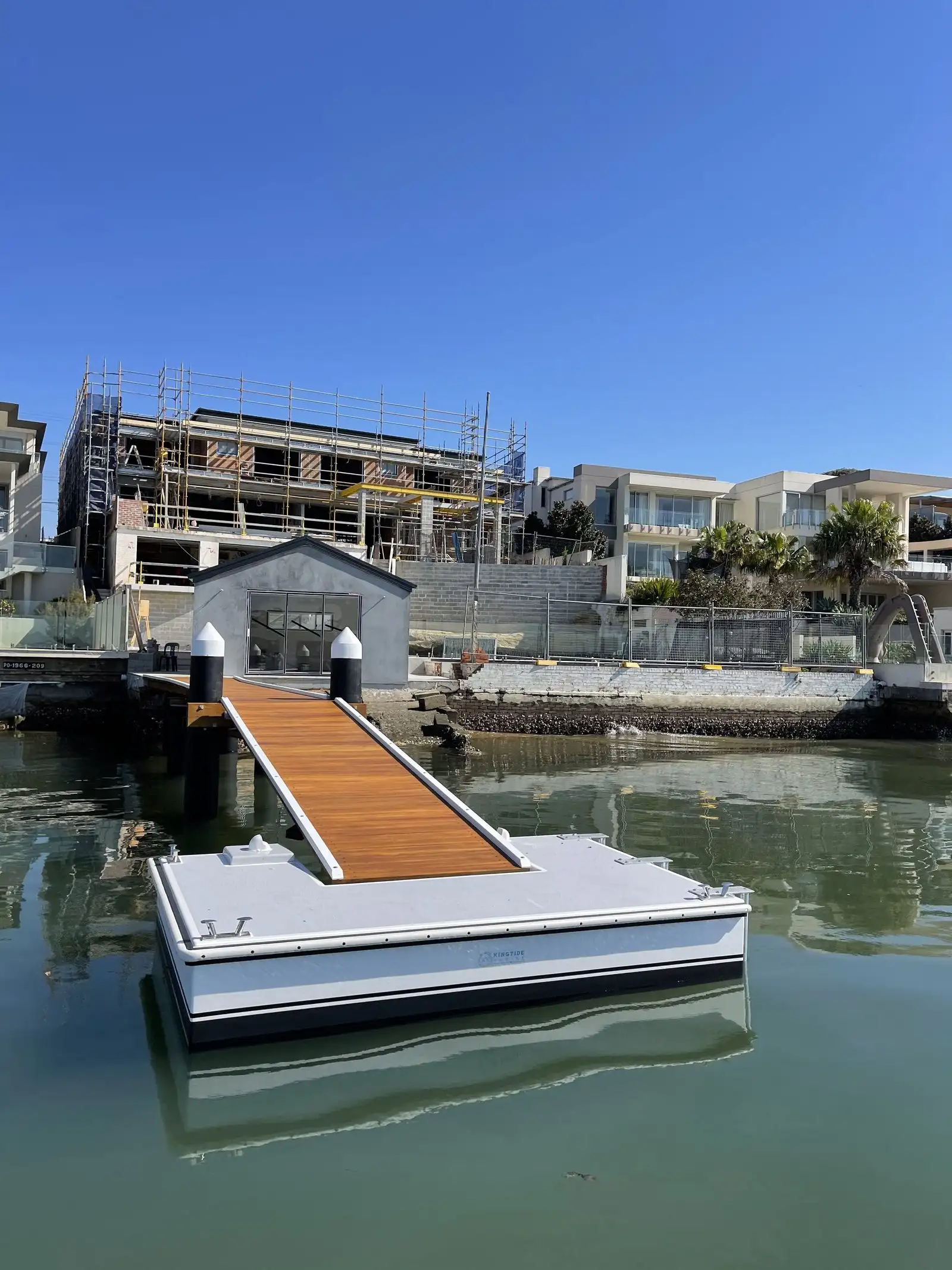 Newly installed Kingtide Marine pontoon and hardwood gangway on flat water in front of a waterfront home under construction