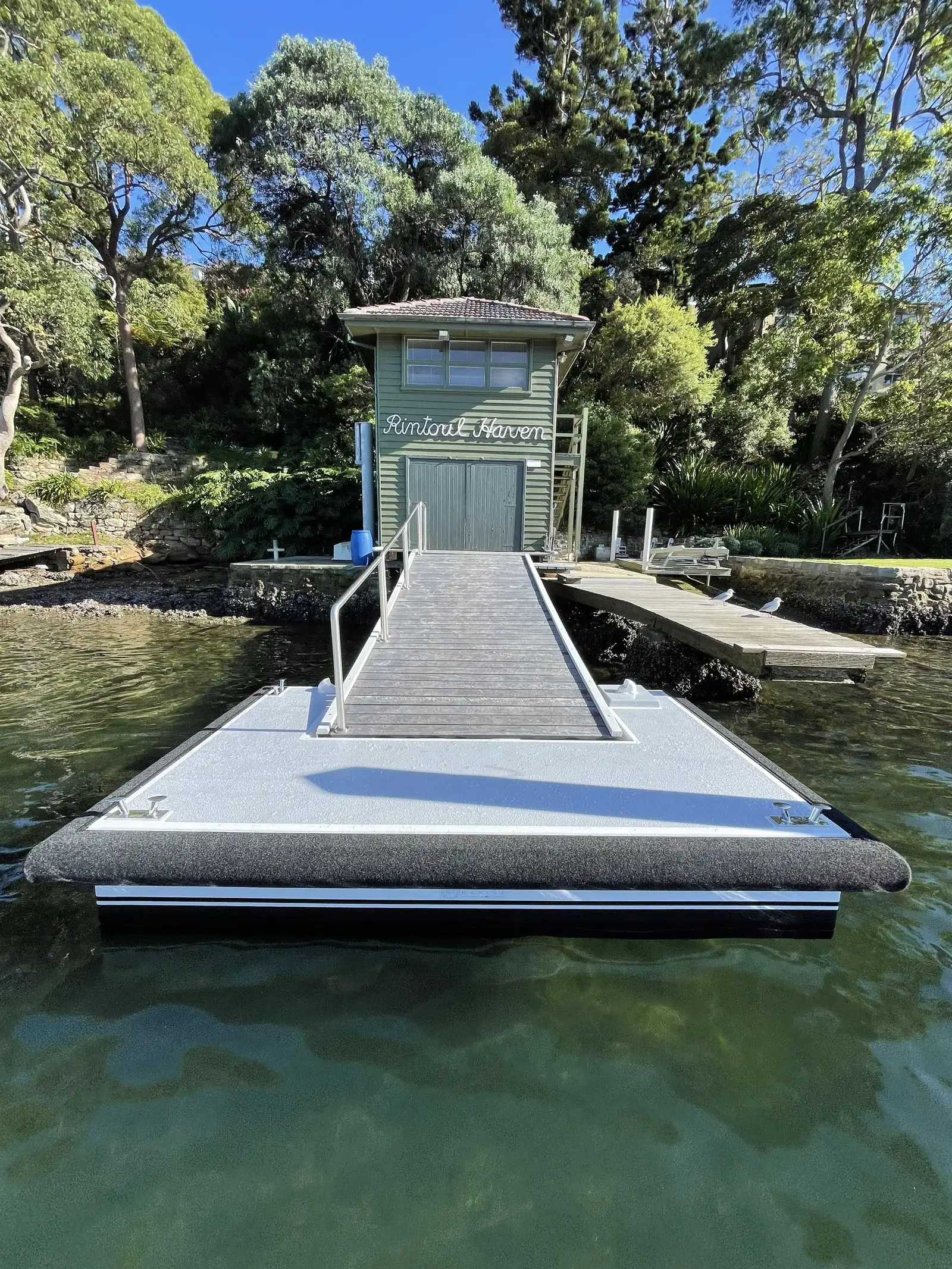 Grey composite-decked pontoon and gangway with a black rubbing strake, moored in front of a green timber boat shed