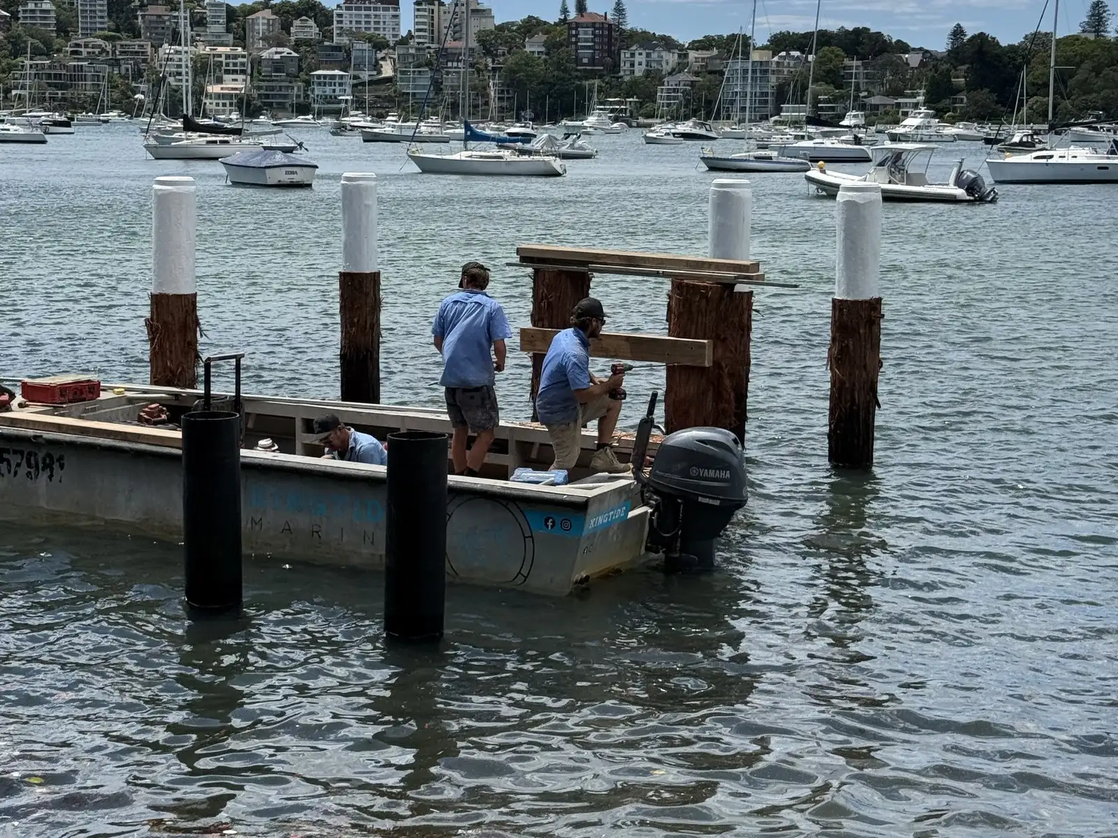 Kingtide Marine crew inspecting and rebuilding jetty piles from the work punt, with a moored fleet behind