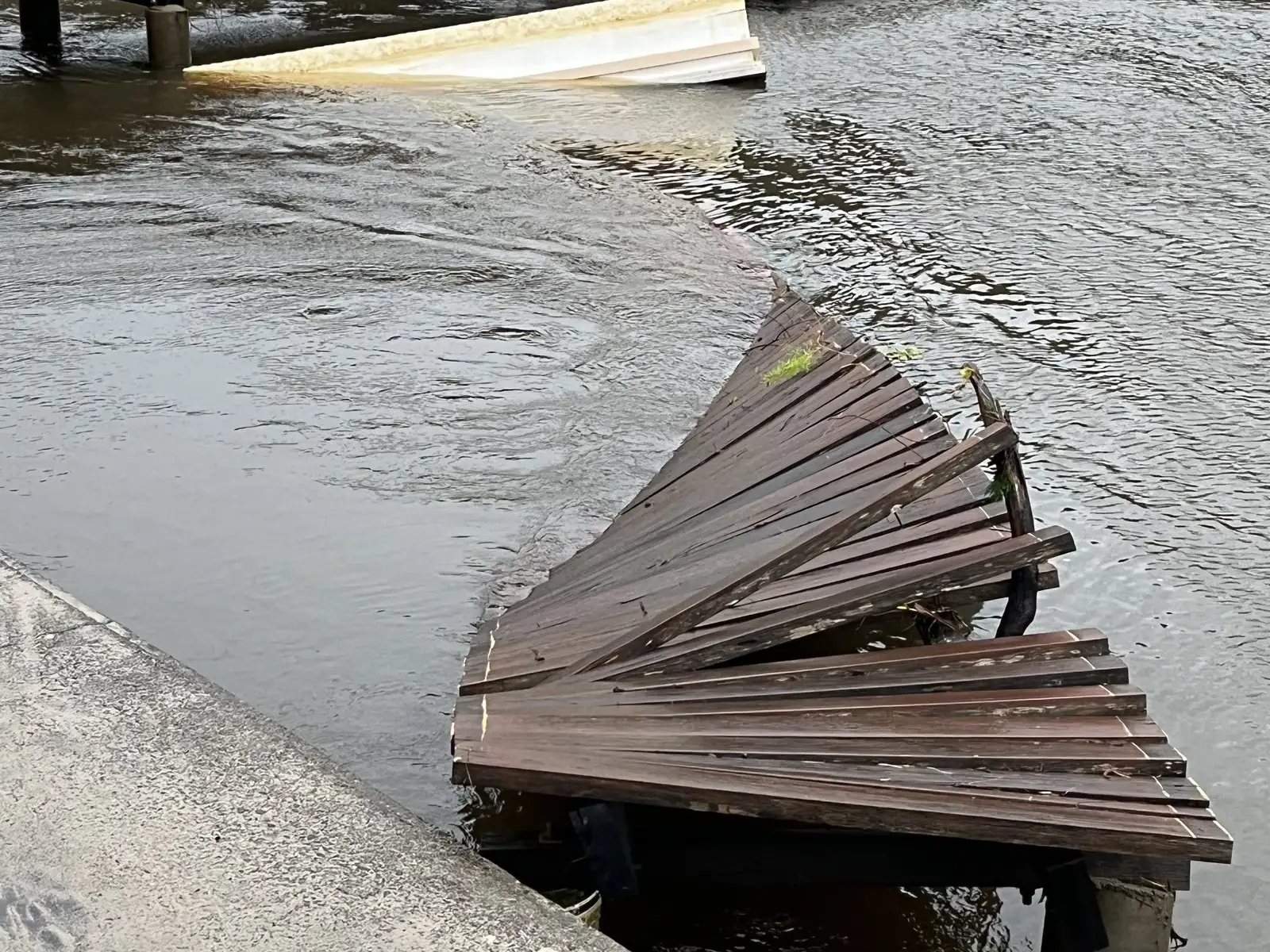 Collapsed timber jetty decking slumped into the water against a seawall, boards twisted out of line