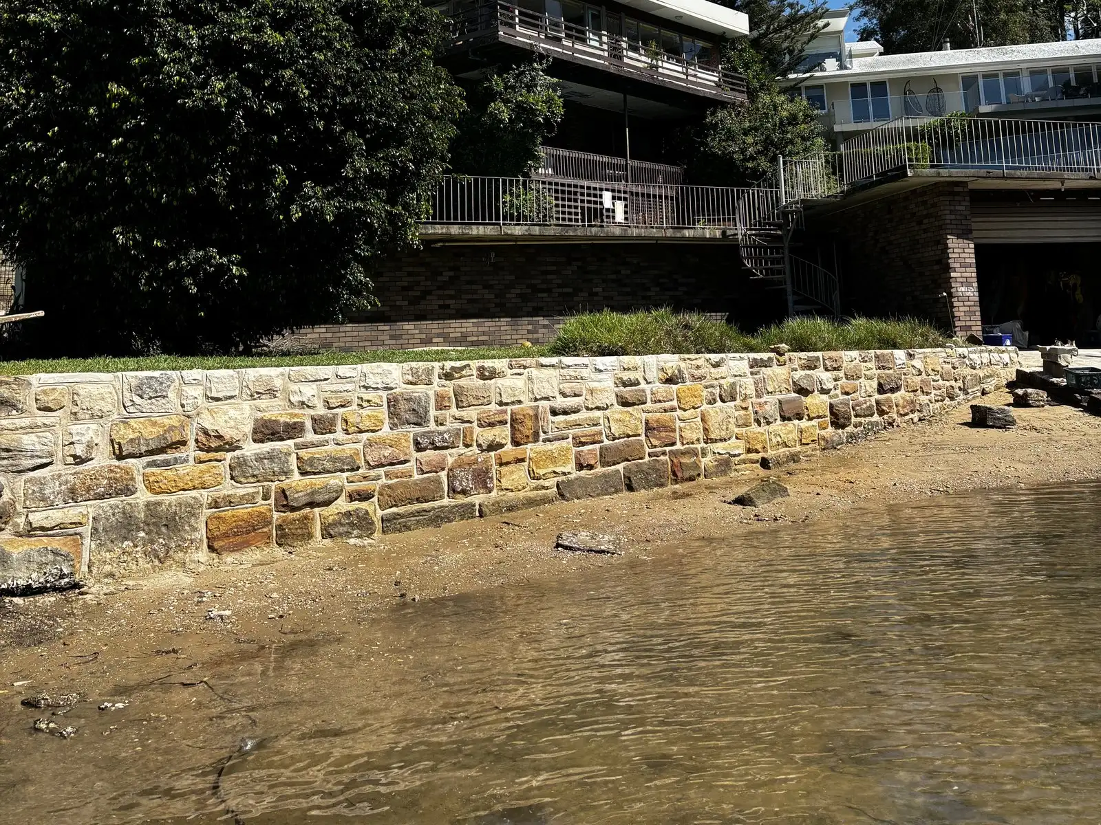A weathered sandstone block seawall holding the lawn above a tidal beach, with the house behind it