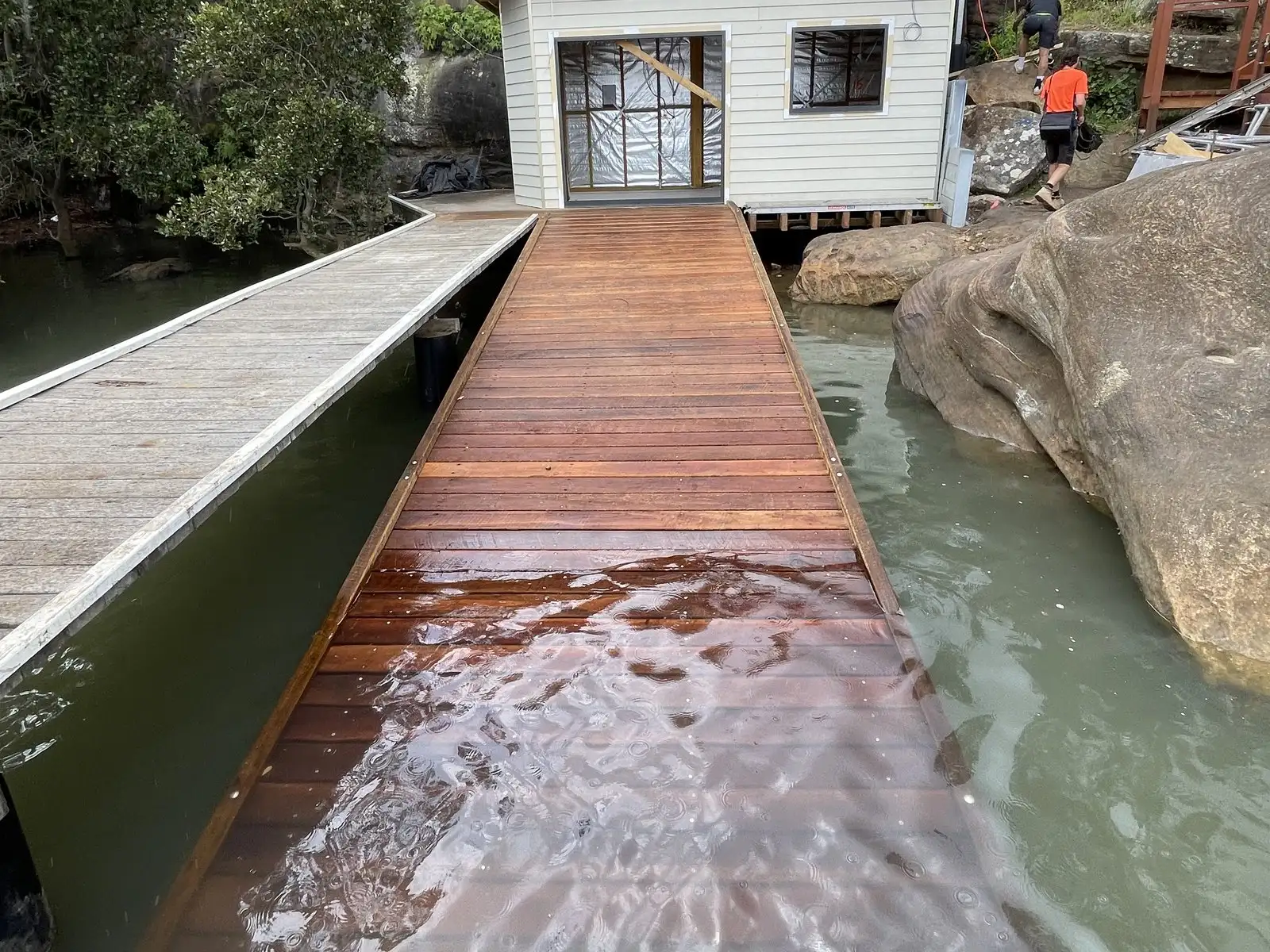 Freshly oiled hardwood skid ramp running down into the water beside a boat shed and sandstone boulders