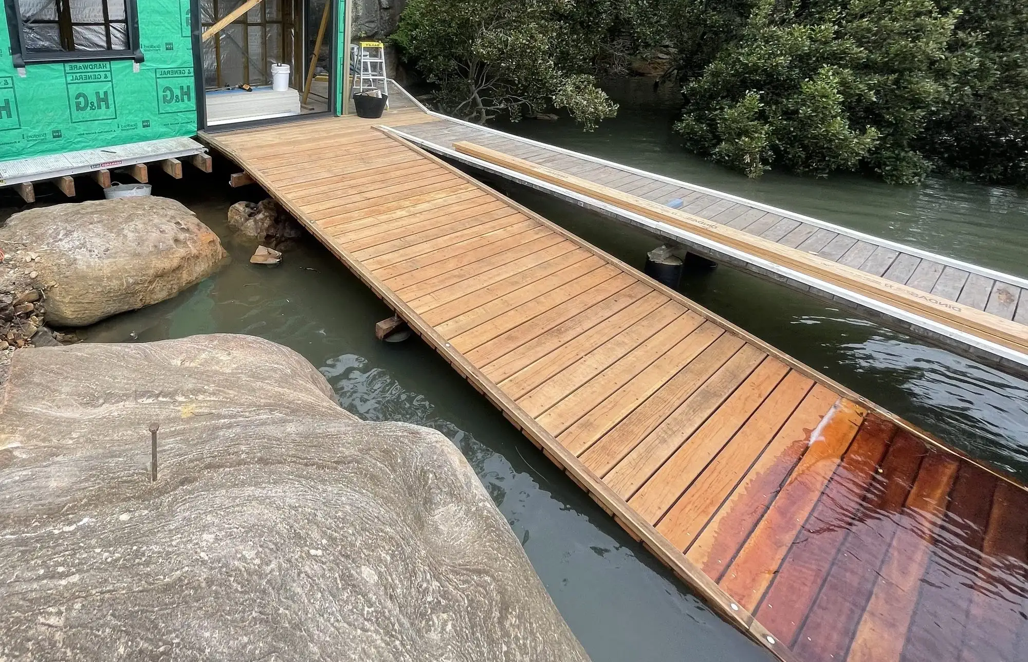 Newly laid hardwood skid ramp running down over the water beside a sandstone boulder, with a boat shed under construction behind