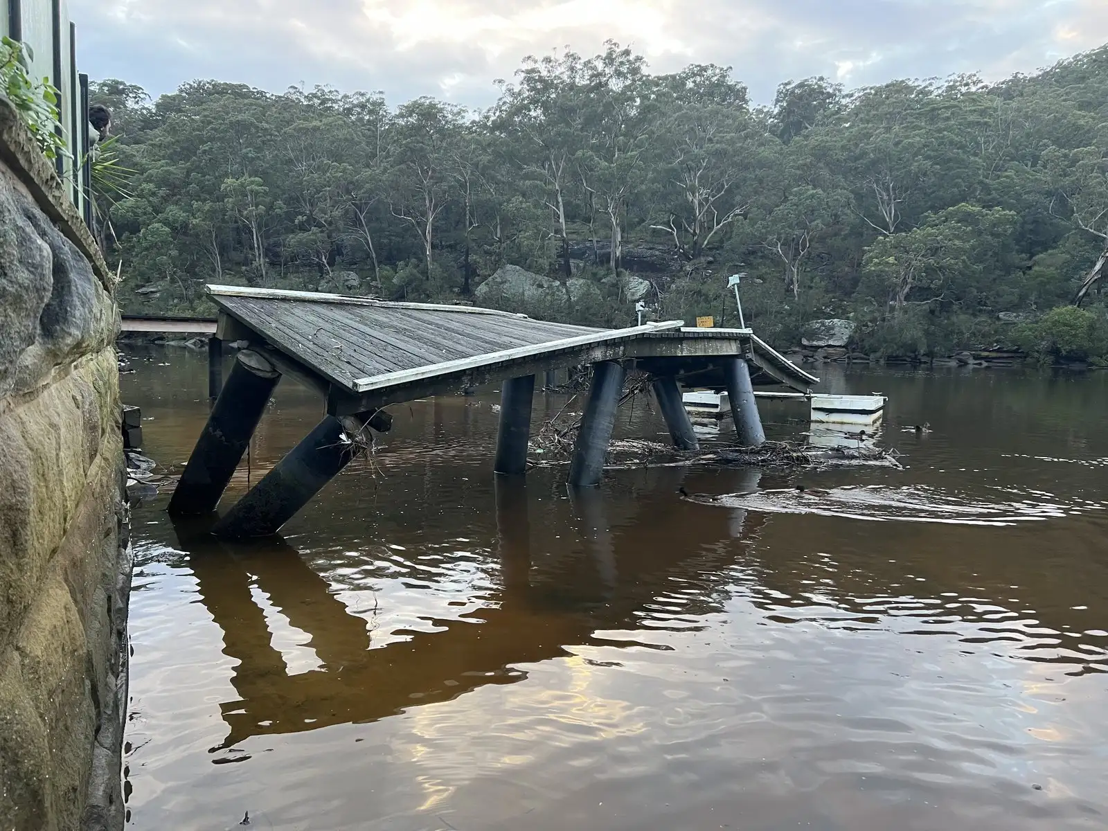 A collapsed jetty deck resting on failed piles in flood water, seen from the foreshore